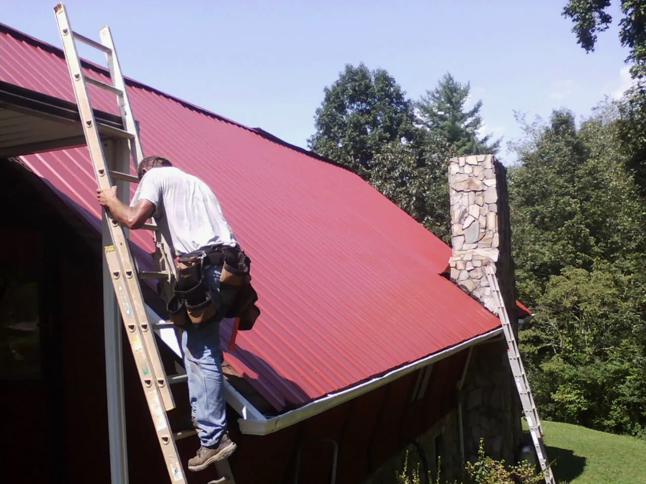 Roofer on a ladder beside a red metal roof and stone chimney — Soffit & Fascia Repair by Roofing Brothers in Great Mills